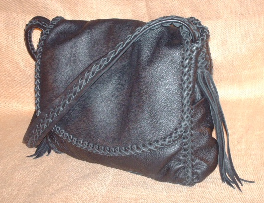  This large Black cowhide leather bag is about 12.5" high by 11" high by 3". It has four pockets - front, inside back, and one on each side. The wide flat leather strap is braided down the center for the length of it. The strap is attached with more braiding that includes long tassels that hang from it. 