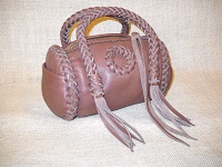  This small Mahogany colored barrel bag has a large brass zipper and is about 7" long with a 5" diameter. It has two 8 strand round braided handles with long tassels hanging from the ends of them. On one side, between the handles, there is a braided circle applique. 