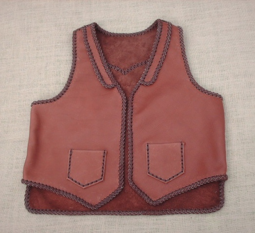  This two tone brown moccasin cowhide leather vest has front lapels, a back pointed yoke, and front patch hip pockets (without flaps). It also has two mated inside patch pockets attached with the same braiding as these outside ones. It also has a 3" back/bottom squared off (not curved) draft flap. 